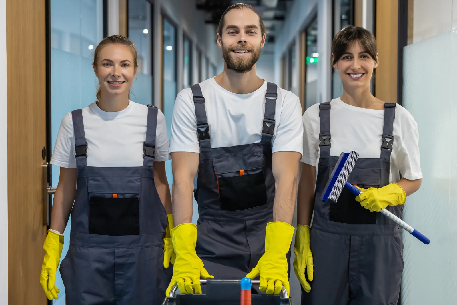 Drei Reinigungskräfte in Arbeitskleidung mit Fensterwischer und Reinigungswagen